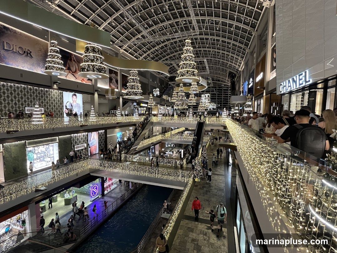 An interior view of a multi-level shopping mall decorated for Christmas. Numerous shops with prominent brand names like Dior, Gucci, and Chanel are visible. Escalators and walkways are filled with shoppers. The mall features a canal or water feature on the lower level and is adorned with festive string lights and Christmas tree-shaped light installations hanging from the ceiling.