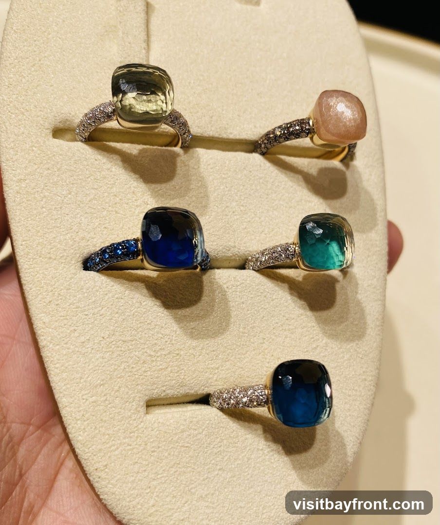 A display of five cushion-cut gemstone rings set in gold bands with diamond accents. The gemstones include a pale green, a peachy pink, a deep blue, a teal, and a dark blue. The rings are presented on a beige display stand.