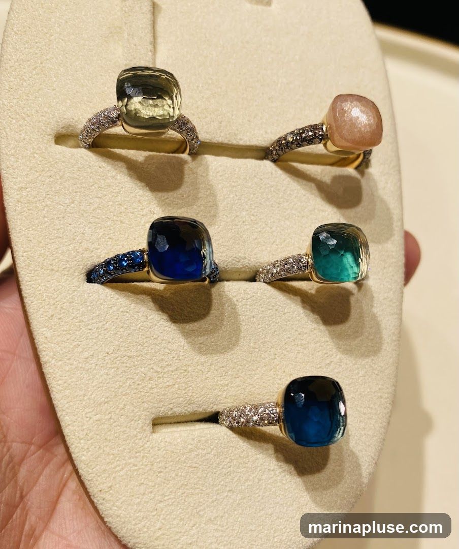 A display of five cushion-cut gemstone rings set in gold bands with diamond accents. The gemstones include a pale green, a peachy pink, a deep blue, a teal, and a dark blue. The rings are presented on a beige display stand.