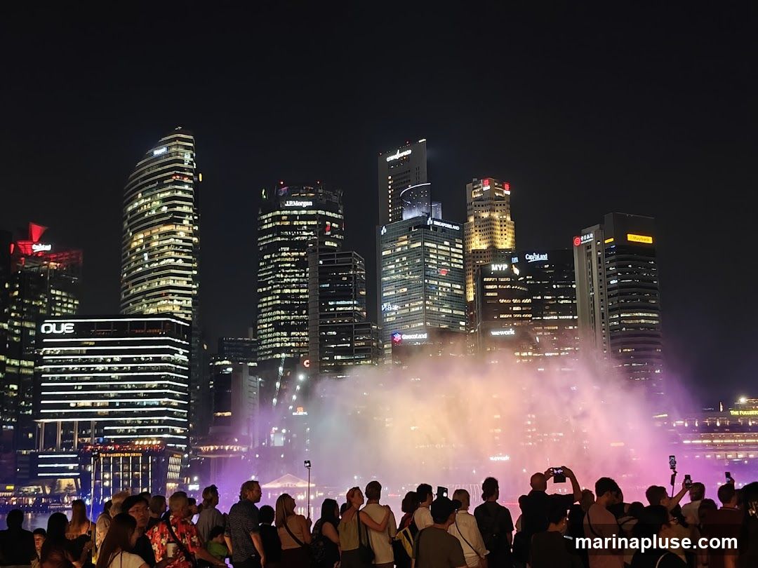 A crowd of people watches a vibrant fountain show at night, with illuminated skyscrapers in the background. The buildings display various company logos, including J.P. Morgan, OUE, and Maybank. The water from the fountain is lit with colorful lights, creating a misty, ethereal effect.