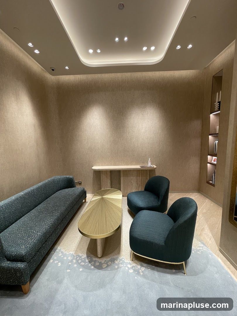 A contemporary lounge area with a textured beige wall, a long dark green sofa, two matching armchairs, and a golden oval coffee table. The floor is covered with a light blue rug featuring a subtle abstract pattern. A narrow console table stands against the wall behind the seating area, and a shelf unit with decorative items is visible on the right. The ceiling has recessed lighting and a modern, curved design.