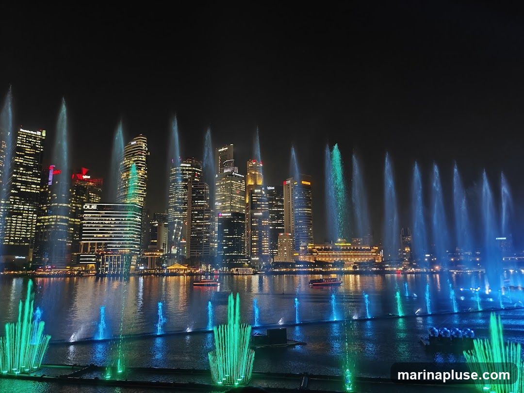 A nighttime view of a city skyline with a vibrant fountain show in the foreground. Tall buildings are illuminated against the dark sky, and numerous water jets, lit with blue and green lights, shoot upwards from the water. Several boats are visible on the water, adding to the lively atmosphere.