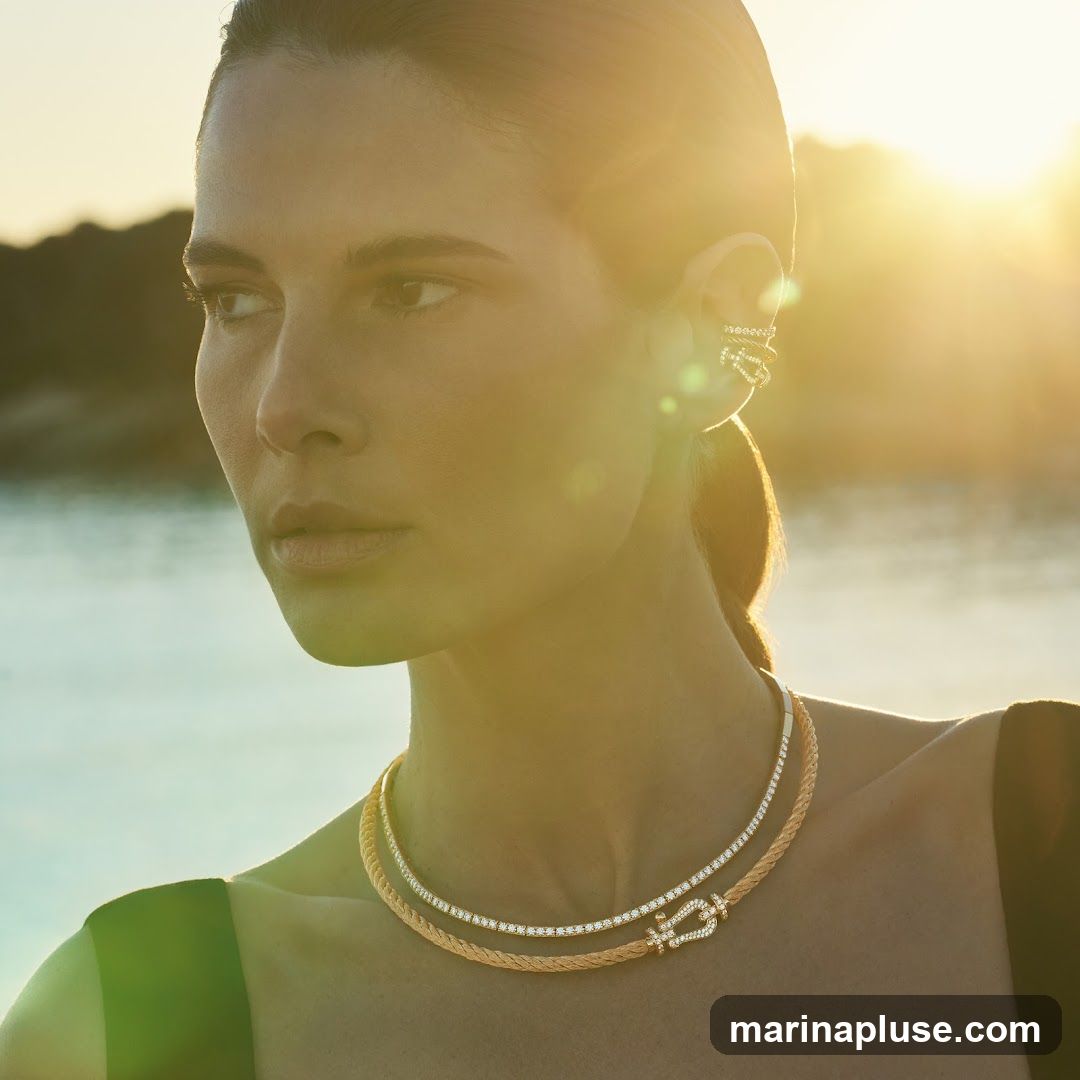 A woman is shown from the chest up, looking to the left, with the sun setting behind her. She is wearing a diamond-encrusted ear cuff and a two-layered necklace. The necklace features a braided gold chain with a diamond-studded clasp and a thinner diamond chain above it. The background is a blurred body of water with a hint of land on the horizon, illuminated by the warm glow of the sunset.