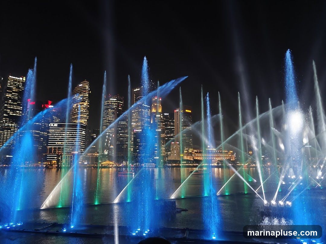 A spectacular nighttime view of a water fountain show with colorful lights, set against a backdrop of illuminated skyscrapers. The water jets are lit in vibrant shades of blue and green, creating a dynamic and mesmerizing display. The city skyline, with its modern architecture, is visible in the background, reflecting on the water.