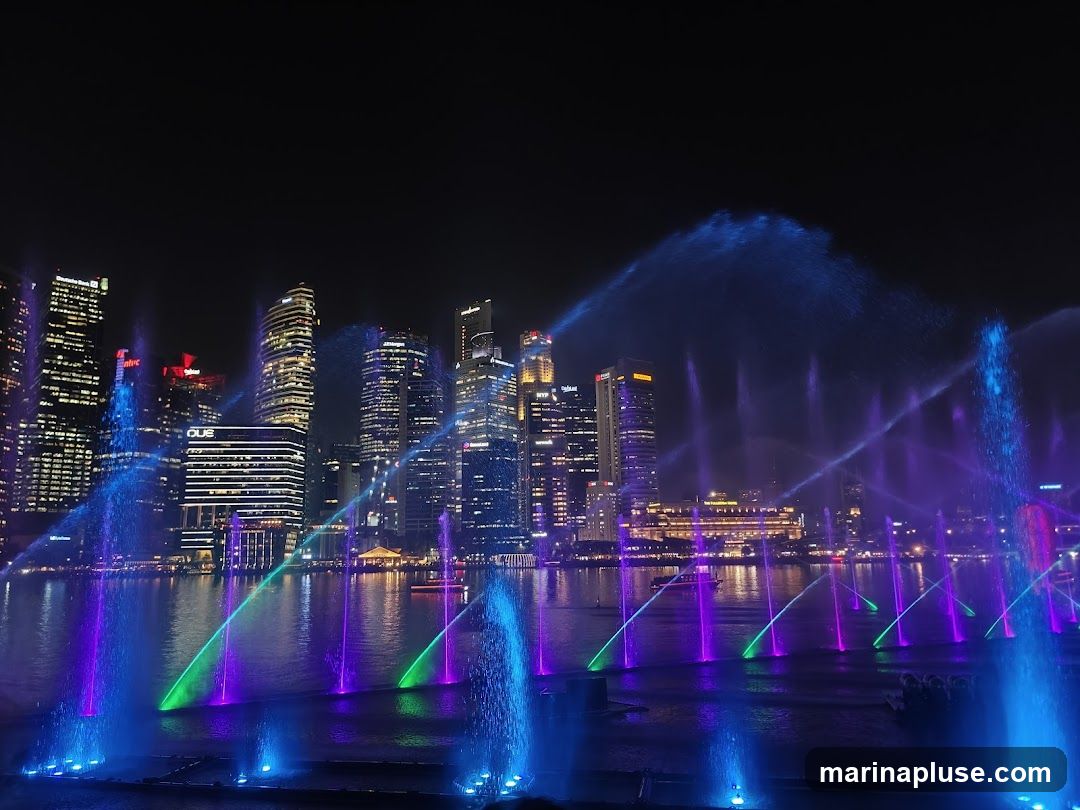 A vibrant nighttime scene featuring a water fountain show with colorful lights in the foreground, set against a backdrop of a modern city skyline. The fountains spray water high into the air, illuminated by blue, purple, and green lights, creating a dynamic and mesmerizing display. The city skyline is composed of numerous tall buildings, some lit up, reflecting on the water. The overall atmosphere is one of urban entertainment and spectacle.