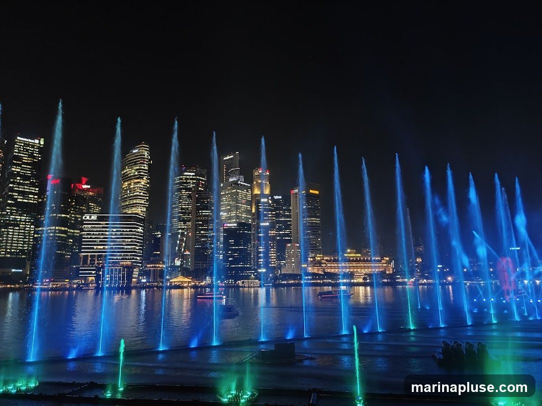 A nighttime view of a city skyline with a large water fountain show in the foreground. The fountains are illuminated with blue and green lights, creating a vibrant display. Tall skyscrapers with illuminated windows form the backdrop, reflecting in the water. Several boats are visible on the water.