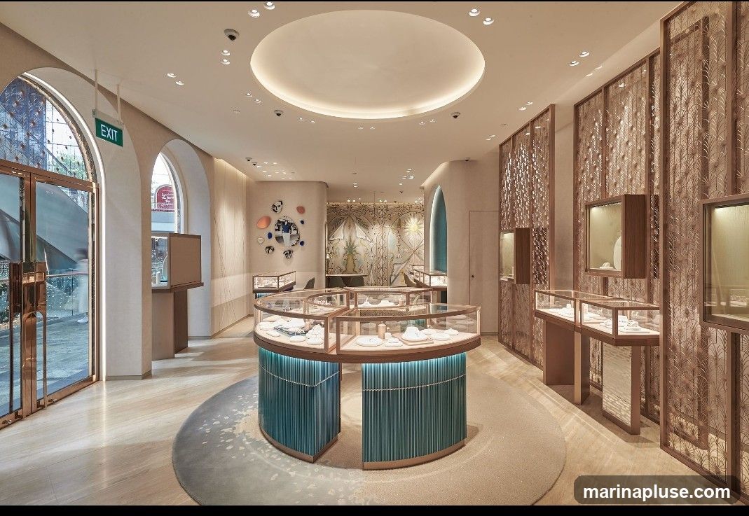 The interior of a high-end jewelry store features elegant display cases filled with jewelry. A central circular display case with teal fluted sides showcases necklaces and rings. Other display cases are arranged along the walls, some integrated into decorative screens. The store has a modern and sophisticated design with soft lighting, arched doorways, and a light-colored marble floor. An "EXIT" sign is visible near the entrance.