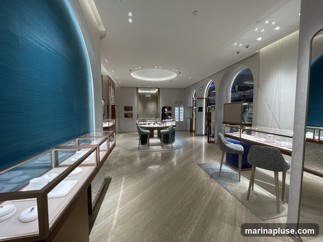 The interior of a modern jewelry store with display cases showcasing necklaces and other jewelry. The store features a large teal textured wall on the left, marble flooring, and elegant lighting. In the background, there are seating areas and more display cases. A person is visible behind a counter in the distance.