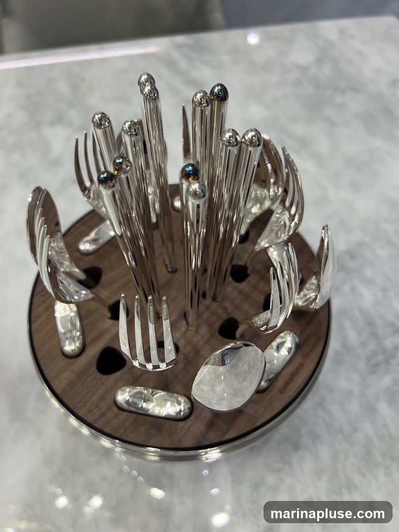 A close-up shot of a polished silverware set arranged in a circular wooden holder. The set includes forks, spoons, and knives, all made of shiny silver. The holder has a dark wood finish with silver trim and appears to be on a marble surface. The brand name 'CHRISTOFLE' is visible on the side of the holder.