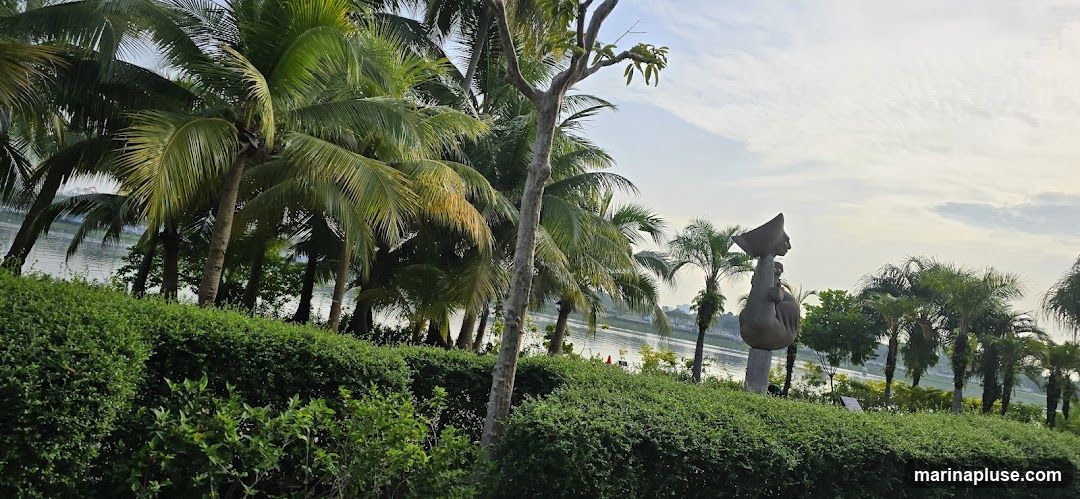 A large, abstract sculpture of a figure carrying a bowl on its head stands in a lush garden setting. The sculpture is positioned on a pedestal and is surrounded by various palm trees and manicured green bushes. In the background, a body of water is visible, with a hazy skyline in the distance. The sky is partly cloudy, with soft light suggesting either early morning or late afternoon.
