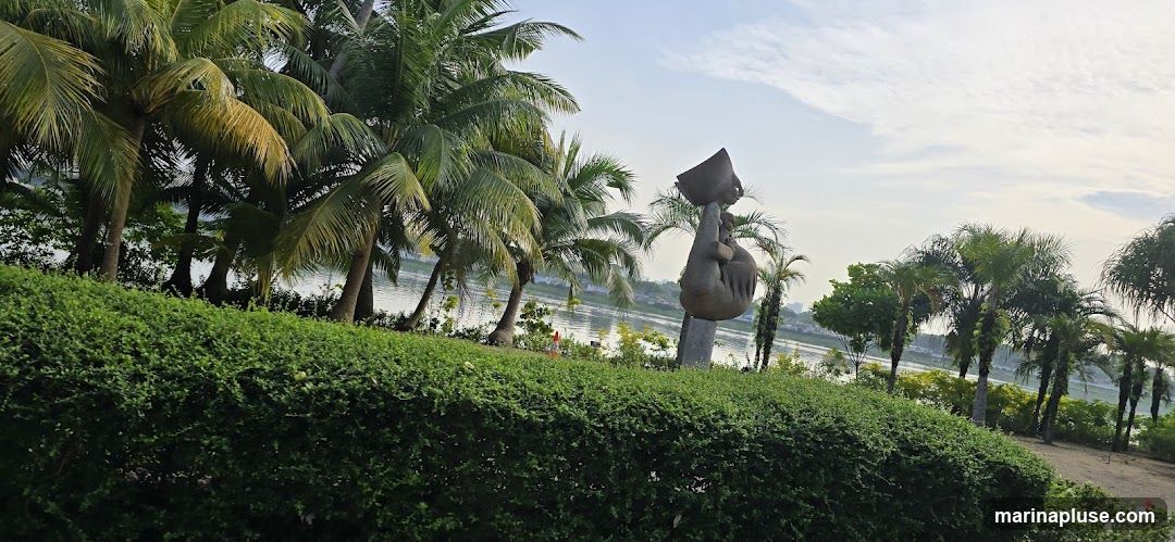 A stone sculpture of a hand holding a bowl stands tall amidst lush greenery and palm trees. The sculpture is positioned near a body of water, with a hazy cityscape visible in the distance. The sky is partly cloudy, suggesting it might be early morning or late afternoon.