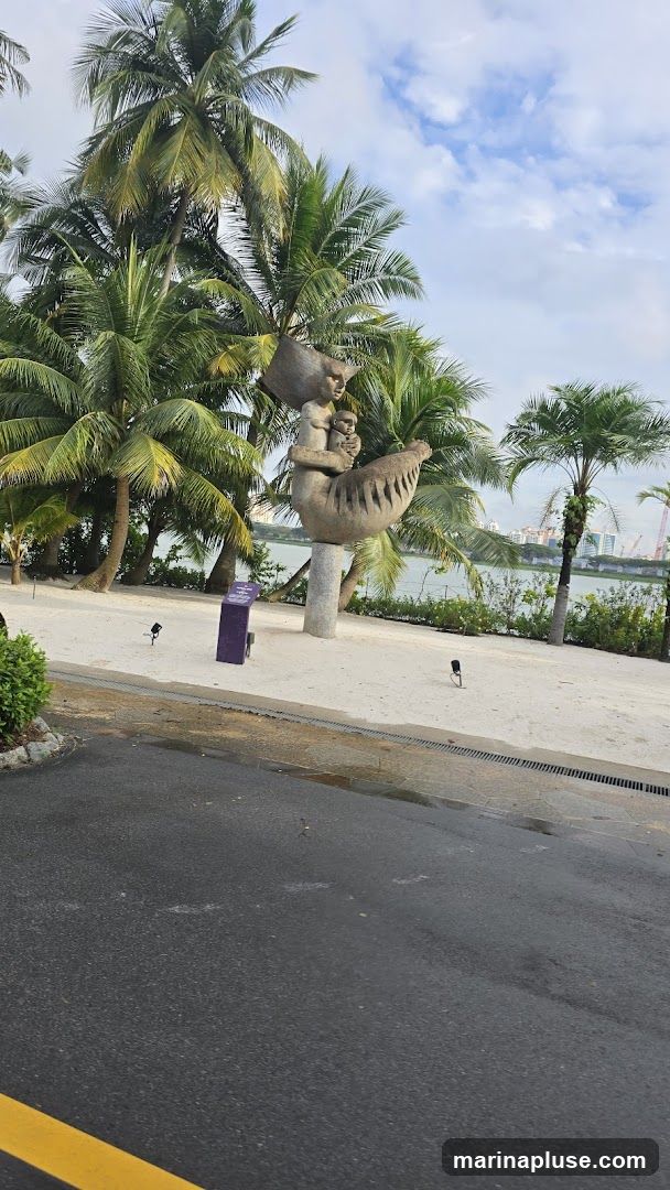 A stone sculpture of a woman holding a child is displayed in a park setting. The sculpture is on a pedestal and surrounded by palm trees and a sandy area. In the background, there is a body of water with a city skyline visible on the horizon. The sky is partly cloudy.