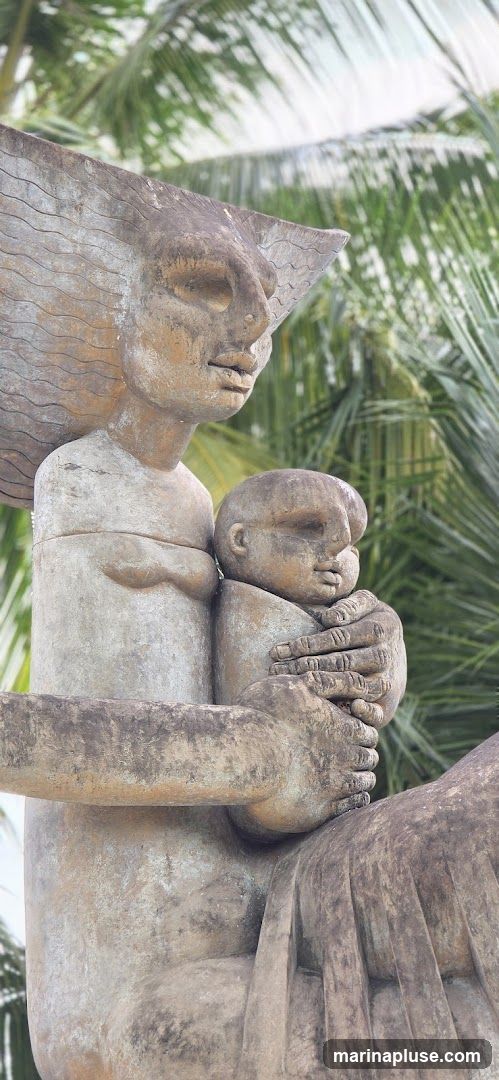 A close-up, eye-level shot of a weathered stone sculpture depicting a mother holding a baby. The sculpture has a rough, textured surface and is set against a backdrop of blurred green palm fronds. The mother's face is serene, with closed eyes and a gentle expression. Her arms cradle the baby, whose face is also rendered with simple, rounded features. The overall impression is one of tenderness and protection.