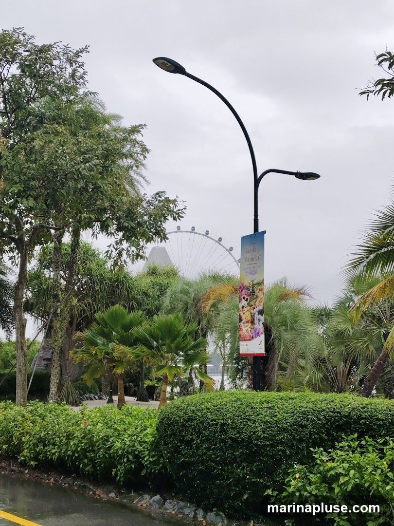 A banner hangs from a lamppost in a lush garden setting. The banner features colorful cartoon characters and the words "Garden Wonders." In the background, a large Ferris wheel is visible against a cloudy sky. The foreground is filled with manicured bushes and palm trees.