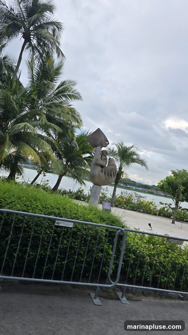 A stone sculpture of a mother and child is displayed near a body of water, surrounded by palm trees and lush greenery. A metal barrier is in the foreground, with a cloudy sky above.