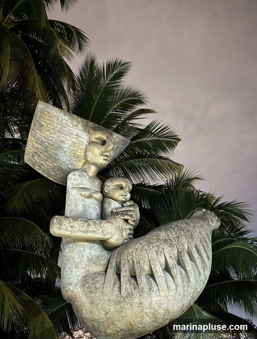 A stone sculpture of a mother holding a baby is set against a backdrop of palm trees and a cloudy night sky. The sculpture has a stylized, abstract design.