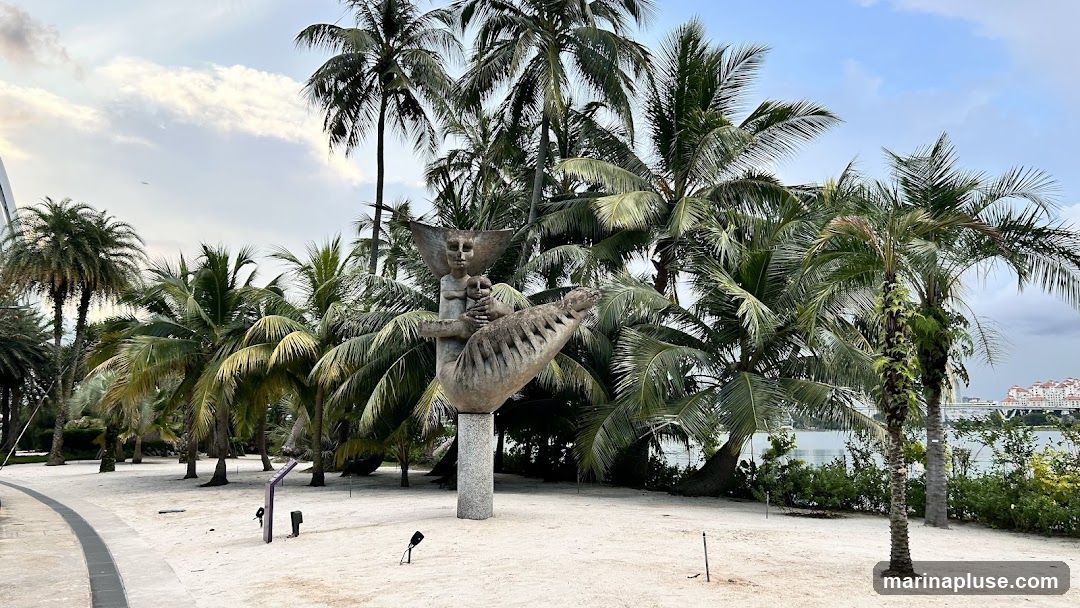 A stone sculpture of a figure holding a child and a creature is displayed in a sandy area surrounded by numerous palm trees. In the background, a body of water is visible with buildings and a bridge on the horizon. The sky is partly cloudy.