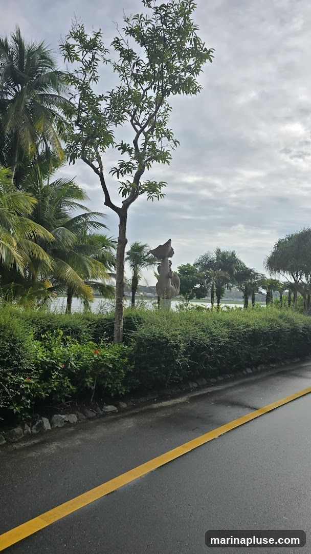 A stone sculpture of a figure holding an object above its head stands amidst lush greenery and palm trees. The sculpture is positioned behind a manicured hedge, with a body of water visible in the background. The sky is overcast. A wet asphalt road with a yellow line runs along the foreground.