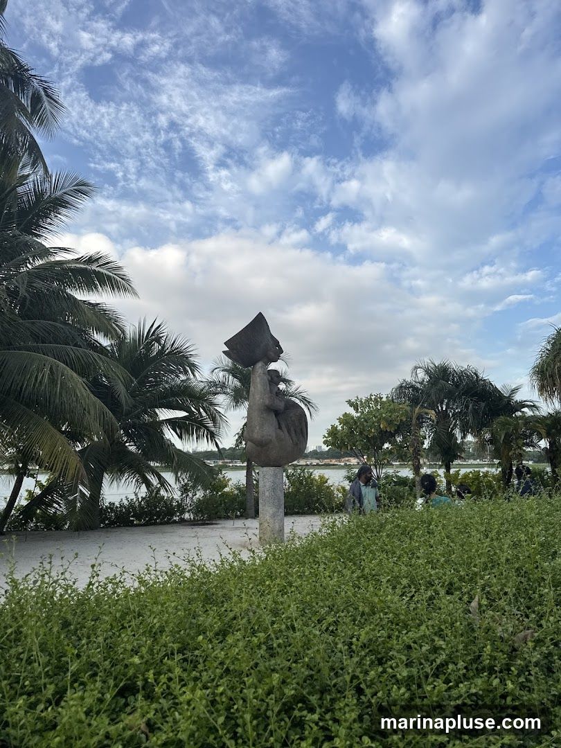A stone sculpture of a figure holding a bowl is situated in a park-like setting. Palm trees and lush green bushes surround the sculpture. In the background, there is a body of water, and a distant cityscape can be seen. Several people are visible in the background, enjoying the outdoor space. The sky is partly cloudy with a mix of blue sky and white clouds.