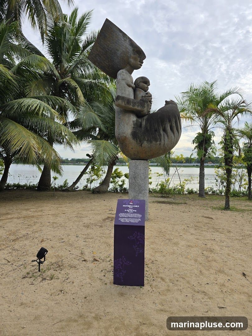 A large abstract sculpture of a mother holding a child is displayed outdoors on a sandy area with palm trees and a body of water in the background. A purple informational sign is placed in front of the sculpture.