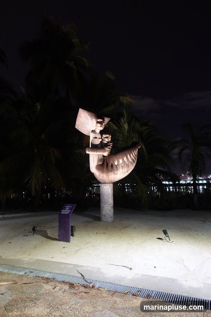 A large, abstract sculpture depicting figures, possibly a mother and child, is illuminated at night. The sculpture is made of a textured, wood-like material and is mounted on a stone pedestal. It is surrounded by palm trees and a sandy ground. In the background, city lights are visible across the water. A small purple informational plaque is placed near the sculpture.