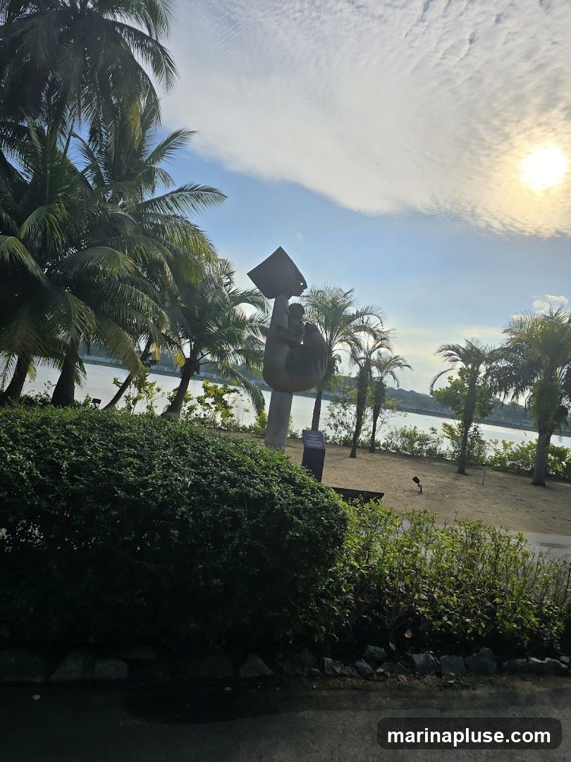 A scenic view of a waterfront park with palm trees and a sculpture. The sun is shining brightly in the sky, partially obscured by clouds. A large, dark green bush is in the foreground, with a paved path behind it. Beyond the bush, a sandy area leads to the water, with more palm trees and a sculpture of a hand holding a bucket. A small sign is visible near the sculpture.