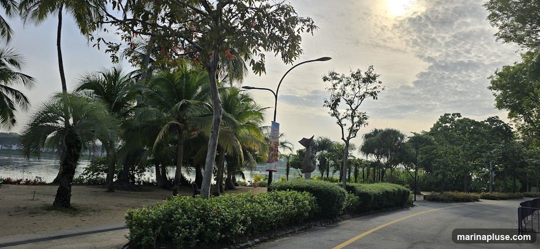 A paved pathway curves through a park-like setting with numerous palm trees and lush green bushes. A modern sculpture, resembling a stylized animal or figure, stands amidst the foliage. In the background, a body of water is visible, reflecting the overcast sky. A lamppost with a banner is also present. The sun is partially visible through the clouds, casting a soft light on the scene.