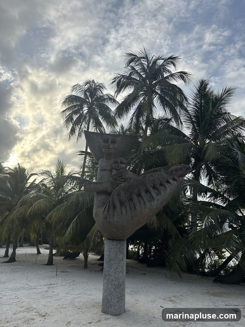A stone sculpture of a figure is displayed outdoors on a sandy area, surrounded by numerous palm trees. The sky above is cloudy with patches of sunlight breaking through. The sculpture is mounted on a tall, grey, granite-like pedestal. The sandy ground appears to be a beach or a similar environment.