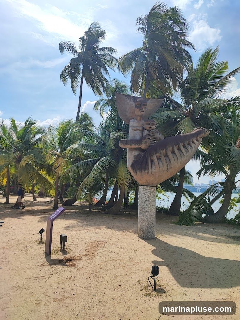 A stone sculpture of two figures, one adult holding a child, is displayed on a sandy beach. The sculpture is mounted on a granite pedestal. Surrounding the sculpture are numerous palm trees, with lush green fronds swaying in the breeze. The sky above is blue with scattered white clouds. In the background, a body of water is visible, with a distant cityscape on the horizon. A purple information plaque stands to the left of the sculpture, with small spotlights placed around the area.