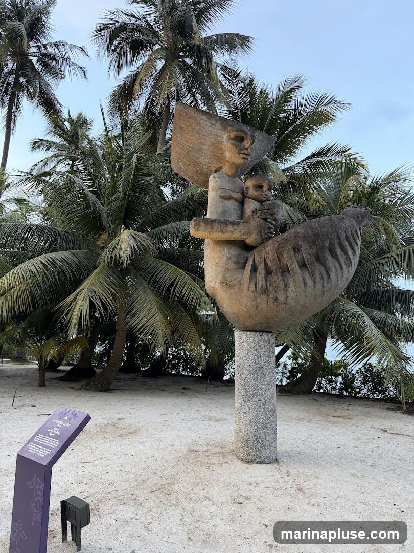 A stone sculpture of a mother holding a child is displayed outdoors on a sandy ground, surrounded by palm trees. A purple informational plaque is visible to the left of the sculpture.