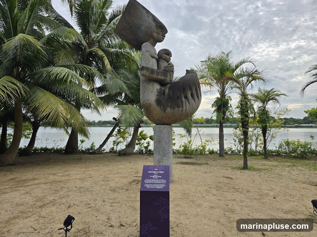 A large stone sculpture titled "Mother & Child" by Dr. Ng Eng Teng stands on a pedestal in a sandy park area. The sculpture depicts a mother holding a child, with an abstract, oversized headpiece. Palm trees surround the sculpture, and a calm body of water is visible in the background under a cloudy sky. A dark purple informational plaque is placed in front of the sculpture's base.