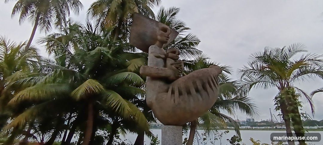 A stone sculpture of a mother holding a child is depicted against a backdrop of lush palm trees and a cloudy sky. The sculpture is abstract in style, with the figures appearing somewhat stylized. In the distance, a body of water and a cityscape are visible.