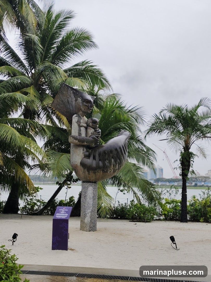 A large, abstract bronze sculpture depicting a mother holding a child, with wings or large leaf-like structures forming a protective enclosure. The sculpture is mounted on a speckled granite pedestal. It is situated outdoors in a sandy area with palm trees and lush greenery in the background. In the distance, a body of water and city skyline with cranes are visible. A purple informational plaque stands to the left of the sculpture.