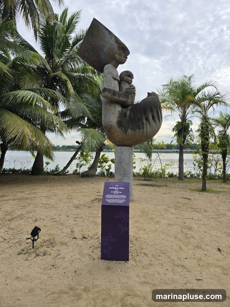A large stone sculpture depicting a mother holding a child is displayed outdoors on a sandy area with palm trees and a body of water in the background. A purple informational sign with floral motifs stands next to the sculpture.