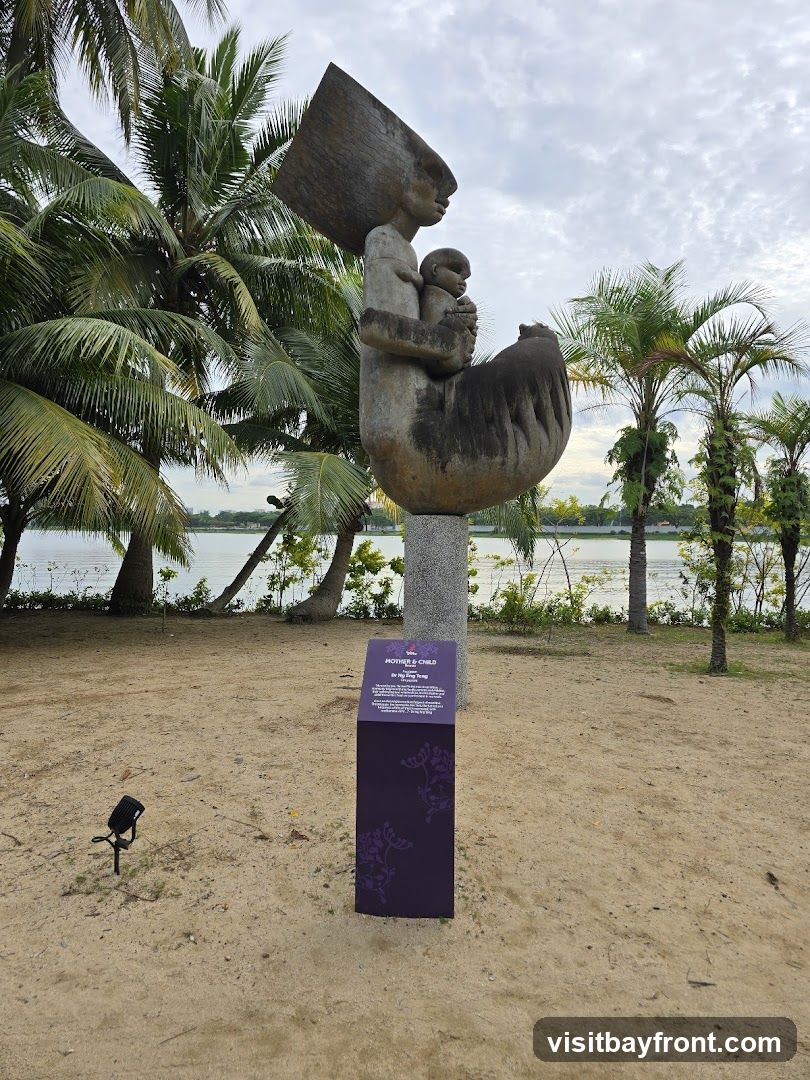 A large stone sculpture depicting a mother holding a child is displayed outdoors on a sandy area with palm trees and a body of water in the background. A purple informational sign with floral motifs stands next to the sculpture.