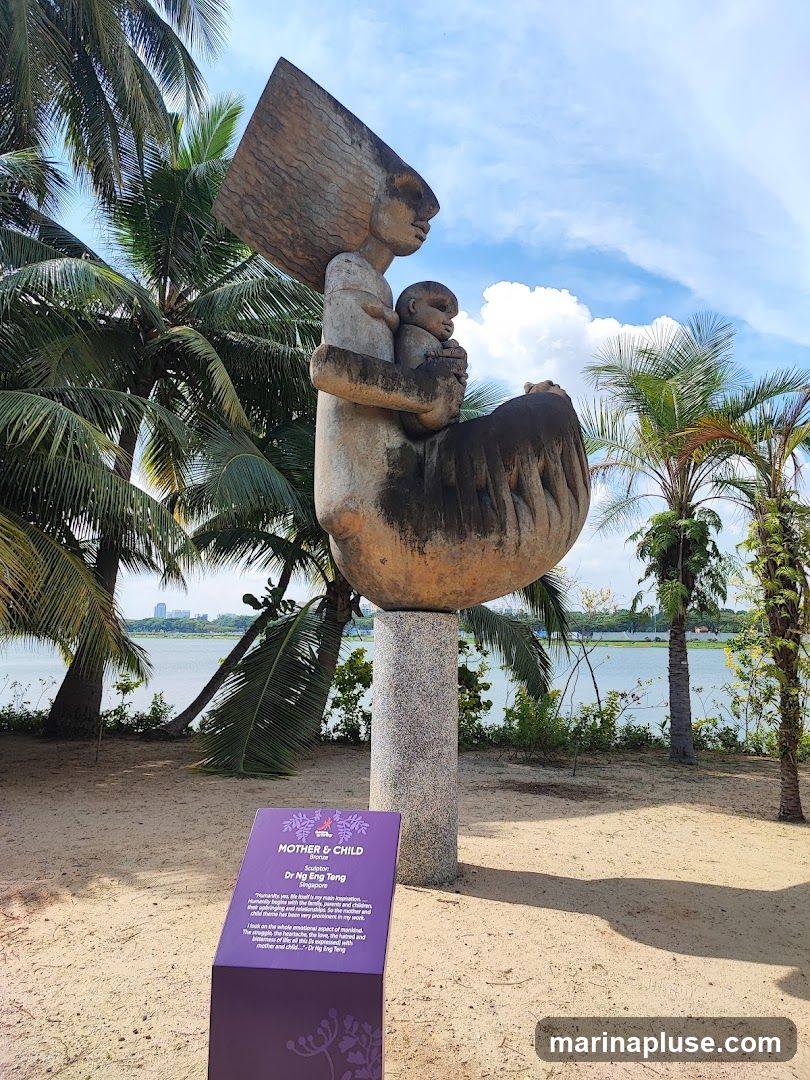 A large abstract sculpture of a mother holding a child stands on a pedestal in a sandy area with palm trees and a body of water in the background. A purple informational plaque is in the foreground.