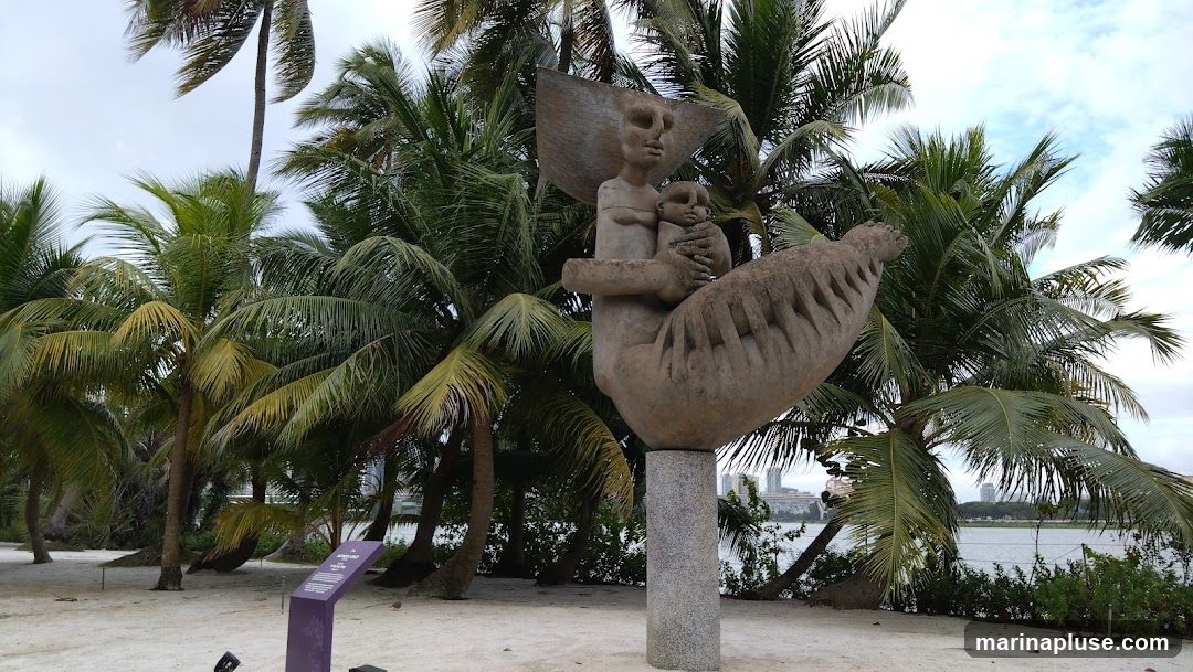 A stone sculpture depicting a mother holding a child is displayed outdoors, surrounded by lush palm trees. The sculpture is mounted on a granite pedestal. In the background, a body of water and distant buildings are visible under a cloudy sky. A purple informational sign is placed to the left of the sculpture.