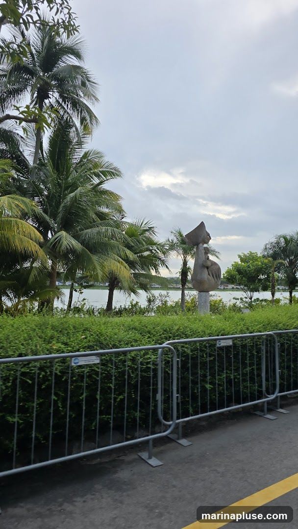 A stone sculpture of a figure holding a large object above its head stands in a park-like setting. It is surrounded by lush green bushes and palm trees. In the background, a body of water is visible, with a cloudy sky overhead. Metal barriers are in the foreground.