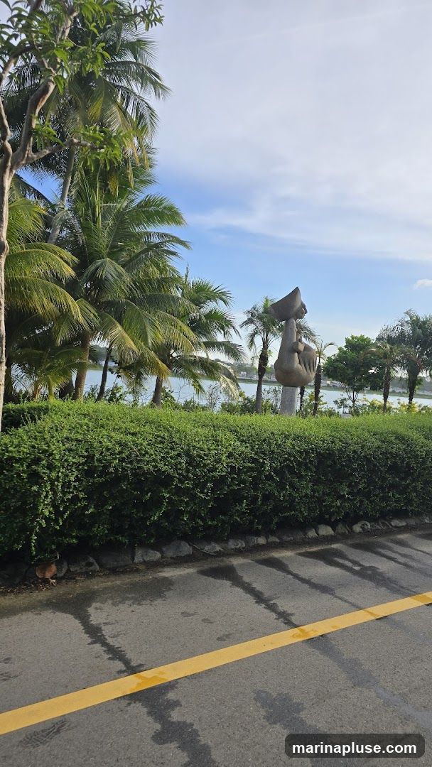 A stone sculpture of a seated figure holding a bowl is visible in the distance, surrounded by palm trees and lush green bushes. The scene overlooks a body of water under a partly cloudy sky. A yellow line is painted on the wet asphalt in the foreground.