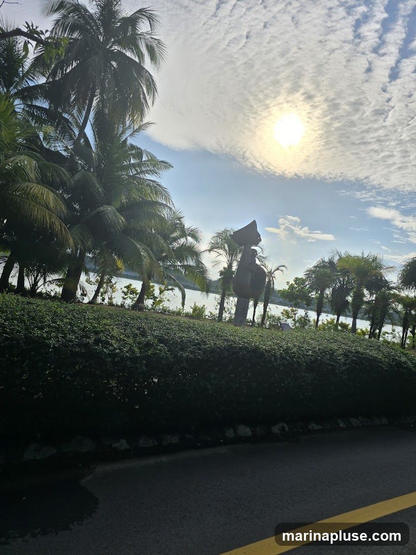A sunny day with scattered clouds over a body of water, lined with palm trees and a manicured hedge. A dark, abstract sculpture stands among the trees. The foreground shows a paved road with a yellow line.