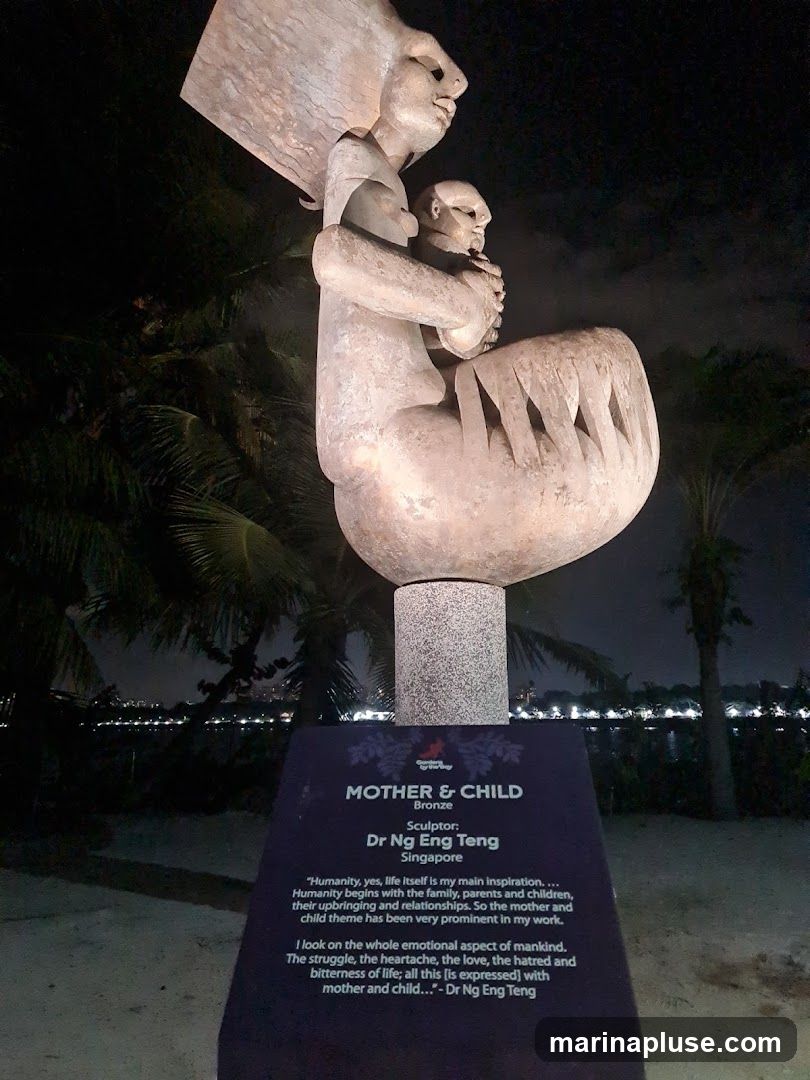 A bronze sculpture titled "Mother & Child" by sculptor Dr Ng Eng Teng of Singapore. The sculpture depicts a mother holding a child. A plaque below the sculpture provides details about the artwork and a quote from the artist. The sculpture is displayed outdoors at night, with palm trees and a waterfront in the background.