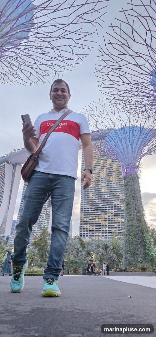 A man stands in the foreground, holding a smartphone and wearing a white polo shirt with a red stripe, blue jeans, and athletic shoes. He is looking at the camera and smiling. In the background, the iconic Supertrees of Gardens by the Bay in Singapore are illuminated, with a tall building visible behind them. The sky is overcast.