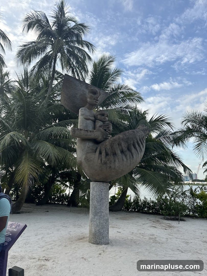 A stone sculpture of a mother holding a child is displayed outdoors on a sandy ground, surrounded by palm trees. The sculpture is mounted on a granite pedestal. In the background, there is a body of water and a distant cityscape with cranes. A sign with text is visible to the left of the sculpture.