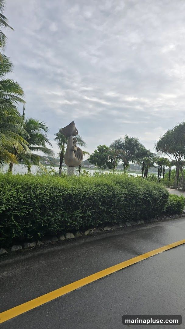 A stone sculpture of a seated figure holding a large object above its head stands on a pedestal. It is surrounded by lush green bushes and palm trees. In the background, a body of water is visible, with trees and buildings on the opposite shore under a cloudy sky. A wet asphalt road with a yellow line runs in the foreground.
