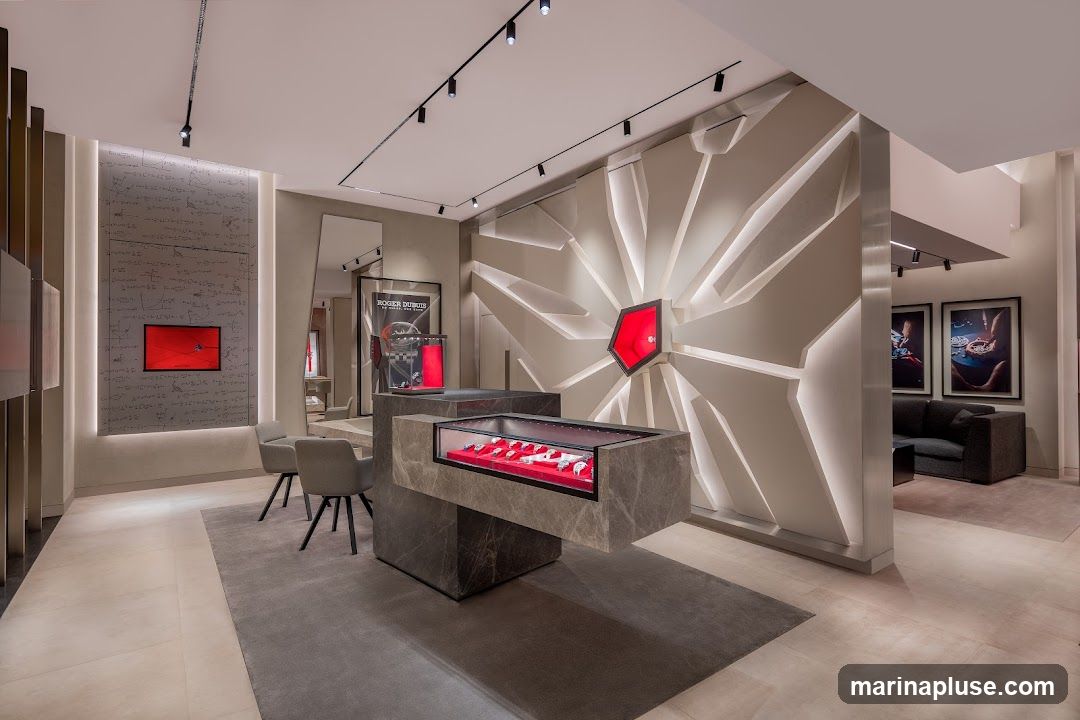 The interior of a modern, high-end watch store. A central display case showcases several watches on red velvet. The back wall features a striking geometric design with a red hexagonal accent displaying a watch. To the left, a wall is covered in what appears to be mathematical equations, with a red screen displaying a watch. Seating areas with chairs and a sofa are visible, along with framed artwork on the walls.