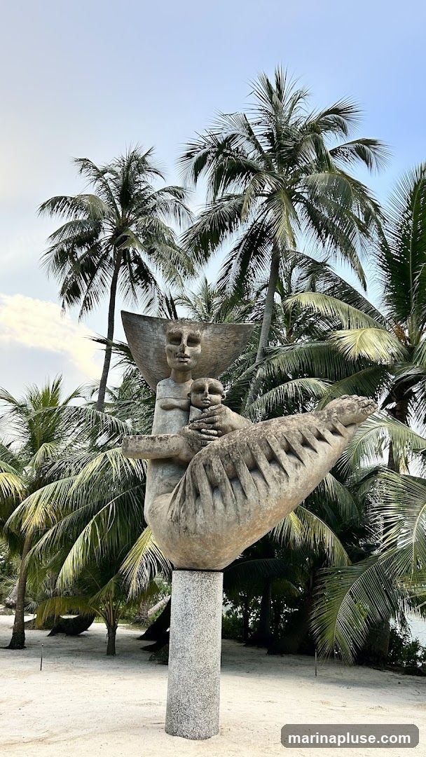 A stone sculpture of a mother holding a child is displayed outdoors, surrounded by lush palm trees. The sculpture is mounted on a granite pedestal. The background features a sandy area and a clear sky with some clouds.