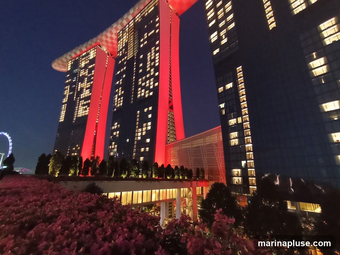 A wide-angle shot of the Marina Bay Sands hotel in Singapore at night. The iconic building is illuminated with red lights, and its windows are lit, creating a striking contrast against the dark blue sky. In the foreground, there is a lush garden with pink flowers and trees. A Ferris wheel is visible in the distance on the left.