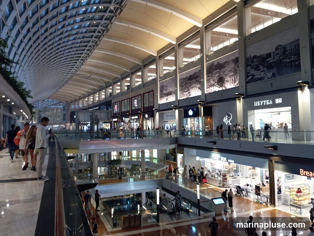 A wide-angle view of the interior of a multi-level shopping mall. The architecture features a curved, lattice-like ceiling and large glass windows. People are seen walking on elevated walkways and escalators. Various shops are visible on different levels, with some displaying signage like 'HEYTEA' and 'BreadTalk'. Large historical murals adorn the upper walls.