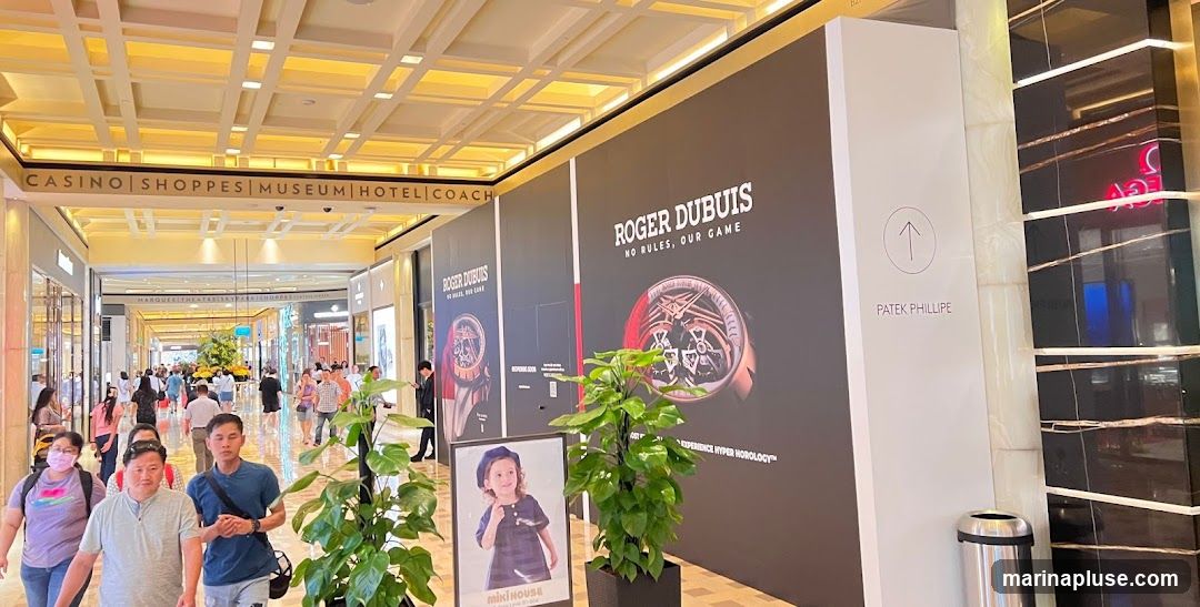 A wide shot of a busy indoor mall walkway. Signs indicate various establishments like CASINO, SHOPPES, MUSEUM, HOTEL, and COACH. Large advertisements for ROGER DUBUIS and PATEK PHILLIPE are prominently displayed on a wall. A smaller sign features a child from MIKI HOUSE. People are walking through the mall.