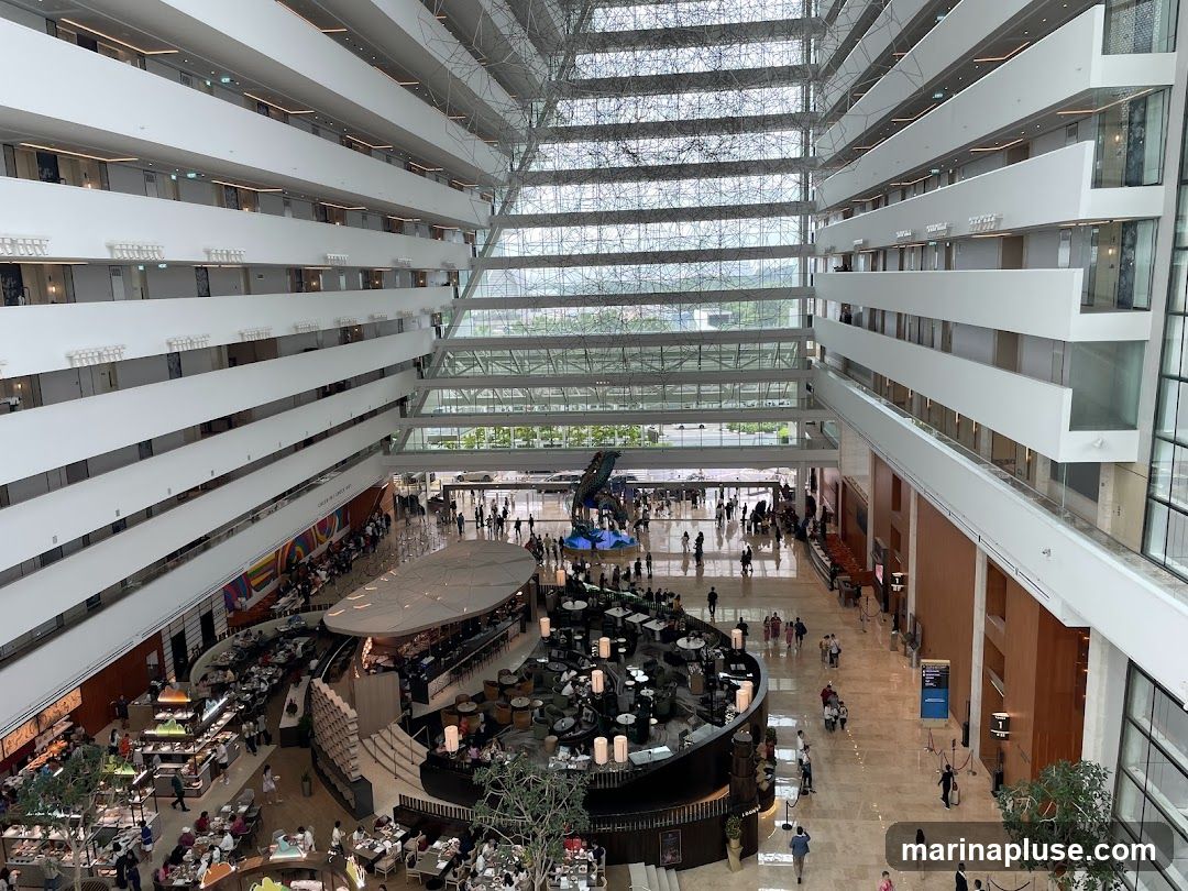 An aerial view of a large, modern hotel atrium. Multiple levels of rooms with balconies overlook a bustling ground floor with seating areas, restaurants, and people walking around. The atrium features a glass ceiling and a distinctive architectural design with white balconies and a central open space.